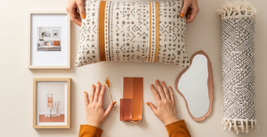 Manos de mujer organizando cojines, marcos de fotos, pintura terracota y espejo en una mesa color crema, decoración económica.