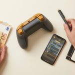 Vista cenital de un controlador de juego, billetes de euro, un cuaderno con 'Ingresos GTA VI' y un smartphone con interfaz de juego.